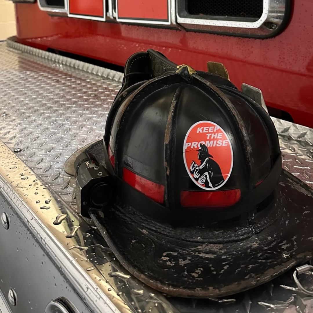 Worn black firefighter helmet with red accents and Keep The Promise sticker rests on wet diamond plate on bumper of ladder truck